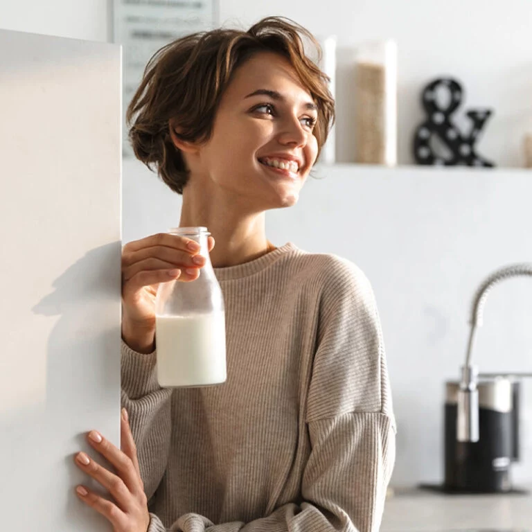 Eine junge Frau mit kurzen braunen Haaren lächelt, während sie eine Glasflasche mit Milch in einer hellen, modernen Küche hält. Sie trägt einen beigen Pullover und steht neben einer weißen Arbeitsplatte.