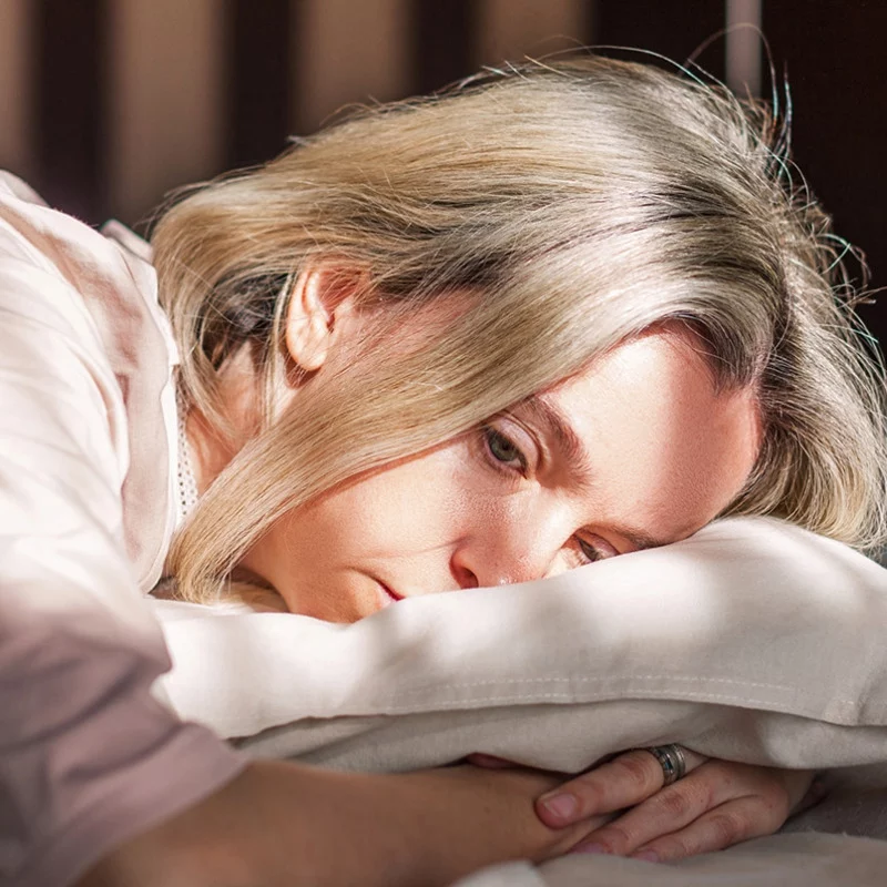 Eine Frau mit blondem Haar liegt auf einem weißen Kissen und sieht nachdenklich und müde aus. Das Sonnenlicht beleuchtet sanft ihr Gesicht, während sie ihren Kopf zur Seite legt, mit einem nachdenklichen Ausdruck und einer Hand, die in der Nähe ihres Gesichts zu sehen ist.