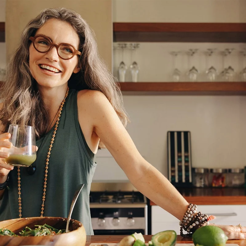 Eine lächelnde Frau mit langen grauen Haaren und Brille steht in einer Küche und hält ein Glas mit grünem Saft in der Hand. Sie trägt ein grünes ärmelloses Oberteil und Perlenketten. Auf dem Tresen vor ihr liegen geschnittene Avocados und eine Salatschüssel.
