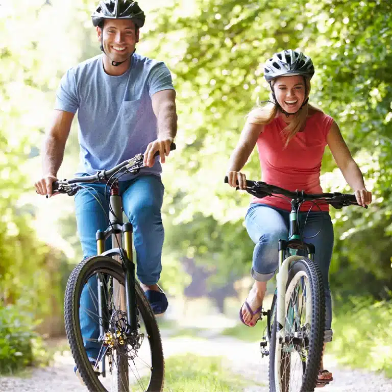 Ein Mann und eine Frau, die Helme tragen, fahren auf einem sonnenbeschienenen, von Bäumen gesäumten Weg Fahrrad, lächeln und genießen gemeinsam eine gemütliche Aktivität im Freien.