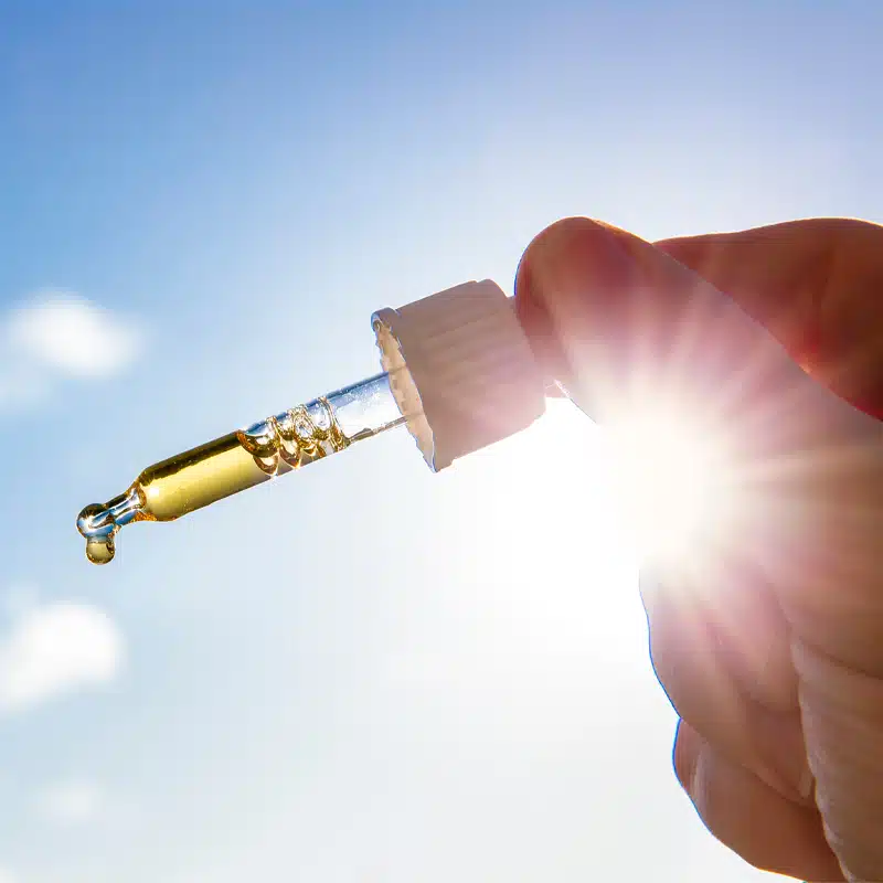 Nahaufnahme einer Hand, die einen mit gelber Flüssigkeit gefüllten Tropfer vor einem hellen, sonnenbeschienenen Himmel hält. Die Flüssigkeit ist im Begriff, einen Tropfen an der Spitze des Tropfers zu bilden.