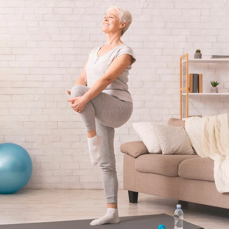 Ältere Frau in grauer Sportkleidung balanciert auf einem Bein und hält ihr Knie an die Brust, während sie lächelt. Sie trainiert in einem gemütlichen Wohnzimmer mit einem Sofa, einer Yogamatte, einer Wasserflasche und einem Gymnastikball in der Nähe.