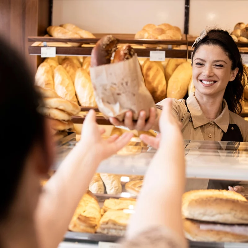 Eine lächelnde Bäckereimitarbeiterin reicht einem Kunden eine Papiertüte mit Brot über den Tresen, während im Hintergrund Regale mit verschiedenen frischen Brotlaiben zu sehen sind.