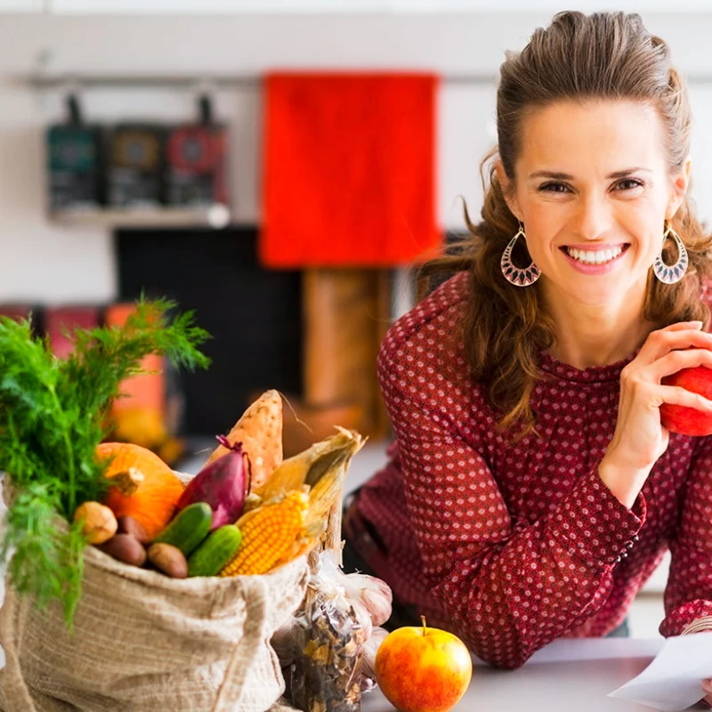 Eine lächelnde Frau in einer rot gemusterten Bluse hält einen roten Apfel in einer hellen Küche. Auf dem Tresen neben ihr steht eine Tüte mit frischem Gemüse und Obst.