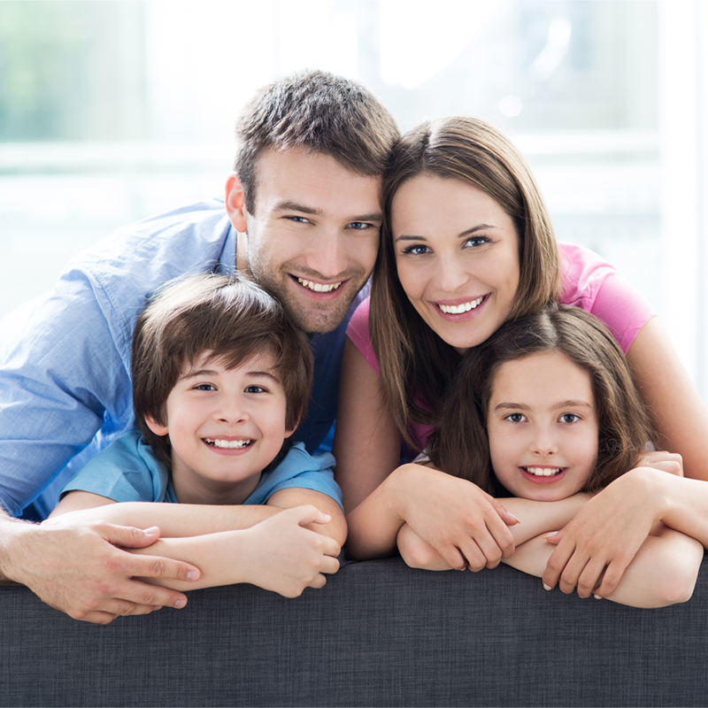 Eine vierköpfige Familie mit Vater, Mutter, einem Jungen und einem Mädchen, lehnt sich über die Rückenlehne einer dunkelgrauen Couch und blickt glücklich direkt in die Kamera. Das Bild dient symbolisch als das biologische Endprodukt unzähliger erfolgreicher Zellteilungen.