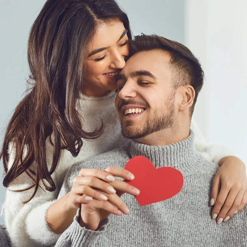 Ein lächelndes Paar in gemütlichen Pullovern umarmt sich, wobei die Frau den Mann von hinten umarmt. Der Mann hält ein rotes Papierherz in der Hand, und beide wirken glücklich und zärtlich.