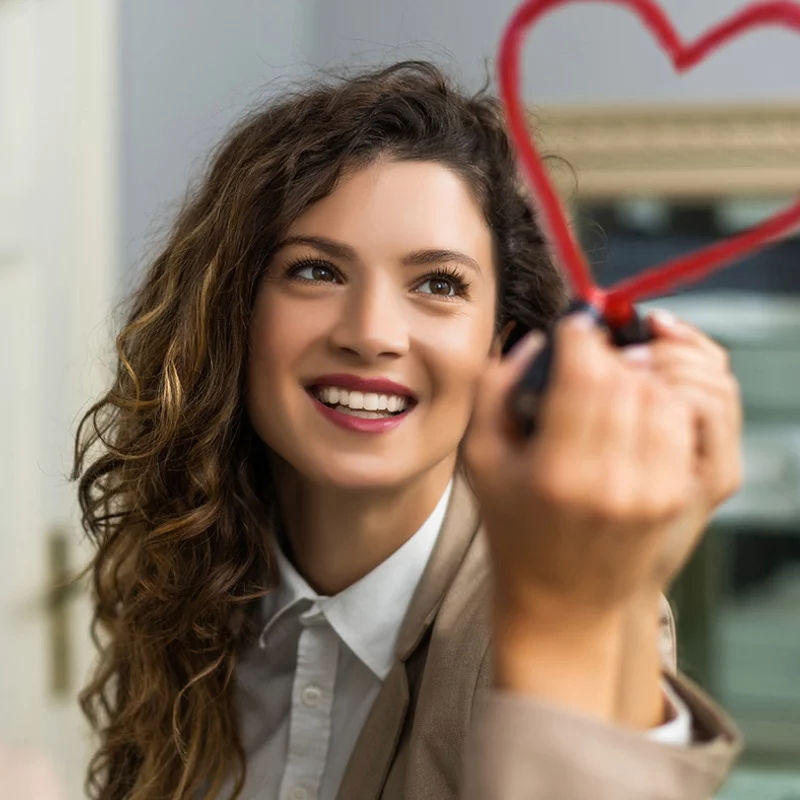 Eine lächelnde Frau mit lockigem Haar zeichnet mit einem Marker ein rotes Herz auf Glas. Sie trägt einen beigen Blazer und ein weißes Hemd und wirkt glücklich und konzentriert auf ihre Zeichnung.