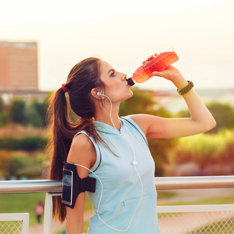 Eine Frau in Sportkleidung trinkt aus einer Flasche, während sie sich im Freien an ein Geländer lehnt. Sie trägt Kopfhörer und ein Smartphone-Armband, im Hintergrund sind städtische Gebäude und Grünanlagen zu sehen.