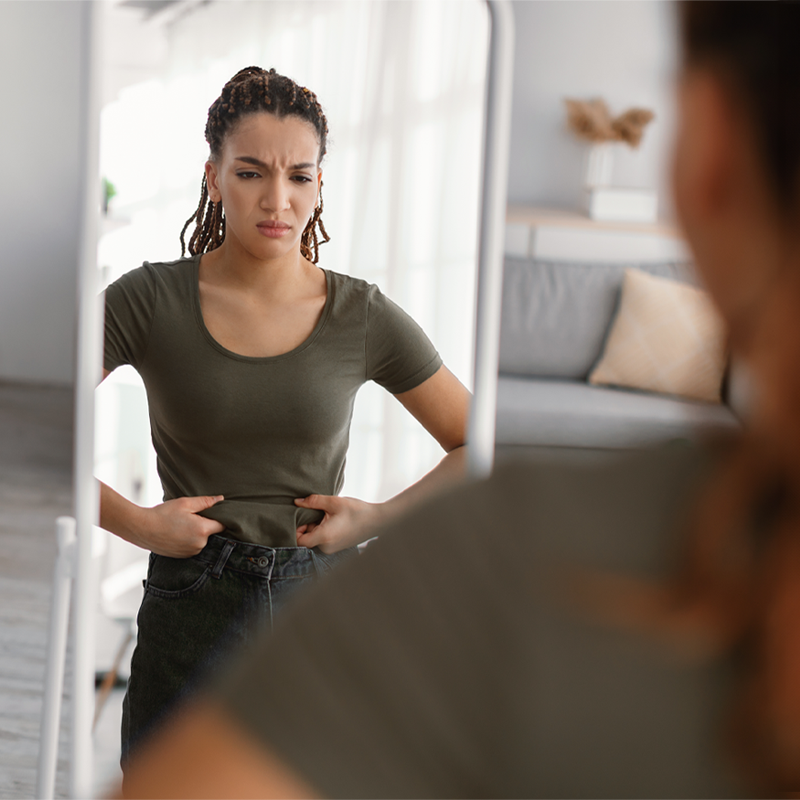 Eine junge Frau steht unzufrieden vor einem großen Standspiegel in einem hell eingerichteten Wohnzimmer mit frustriertem Blick auf ihr Spiegelbild. Sie greift sich mit beiden Händen an ihren Bauch, um vermeintliche Problemzonen zu prüfen.