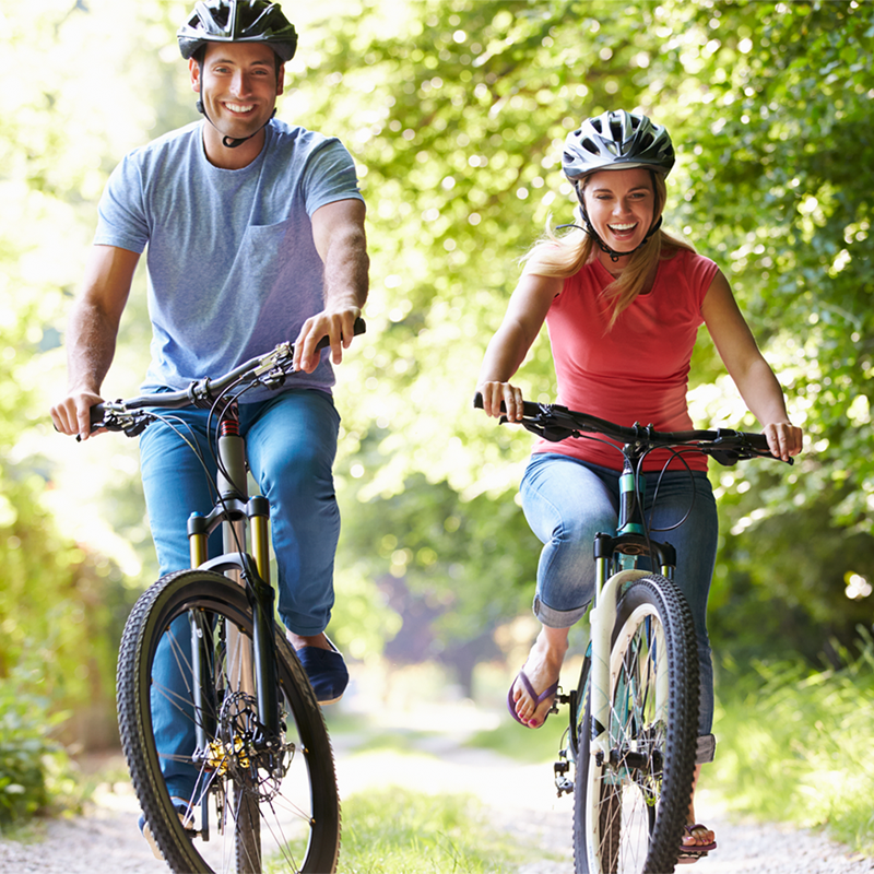 Ein lächelnder Mann und eine Frau, beide mit Fahrradhelm, fahren gemeinsam auf einem sonnigen Waldweg Fahrrad