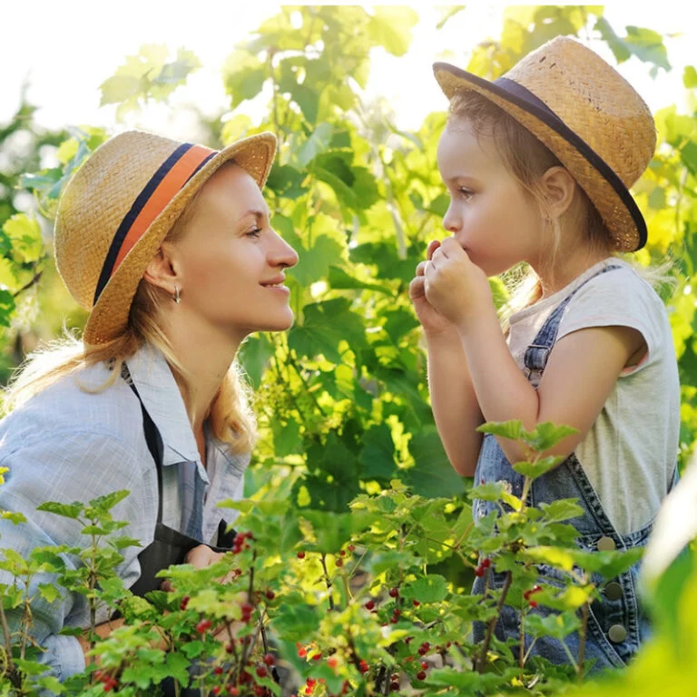 Eine Frau und ein junges Mädchen mit Strohhüten stehen in einem üppigen Garten. Die Frau lächelt das Mädchen an, das Beeren isst und einen Jeans-Overall trägt. Das Sonnenlicht fällt durch die grünen Blätter um sie herum.