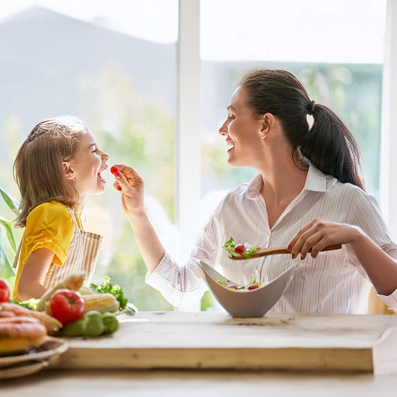 Eine lächelnde Frau füttert ein junges Mädchen mit einer Kirschtomate, während sie an einem Tisch mit frischem Gemüse und einer Salatschüssel in einer sonnenbeschienenen Küche sitzen.