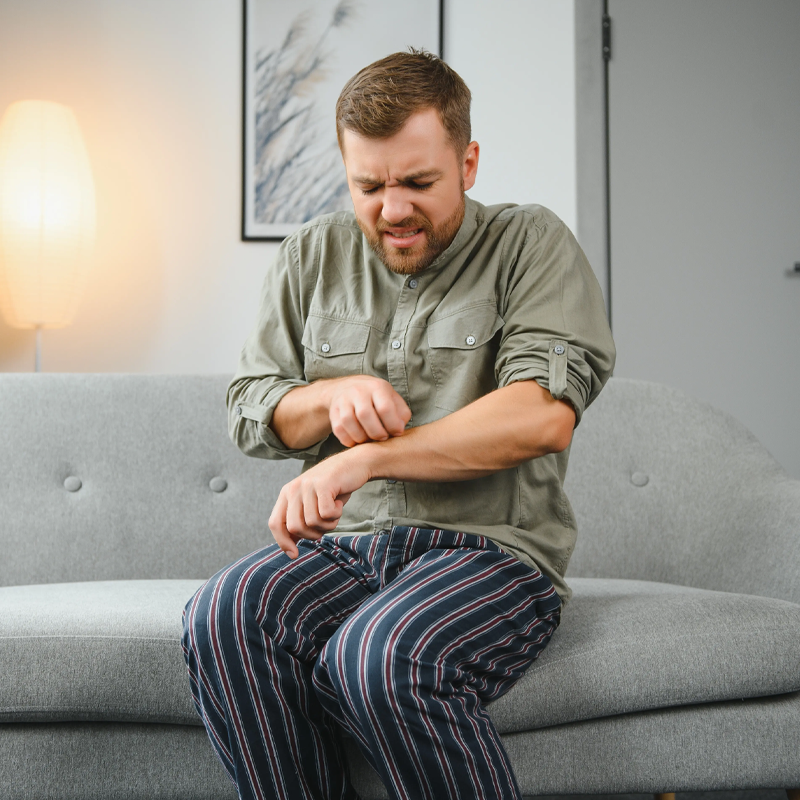 Ein Mann im Schlafanzug sitzt auf einer grauen Couch in einem Haus, kratzt sich am Arm und scheint sich unwohl zu fühlen, möglicherweise wegen eines Juckreizes oder einer Hautreizung. Im Hintergrund sind eine Lampe und ein gerahmtes Bild zu sehen.