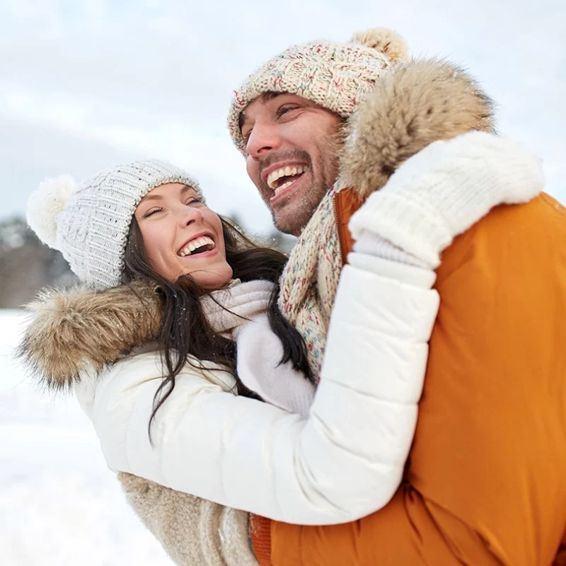 Ein lächelndes, mit Winterjacken, Mützen und Schals bekleidetes Paar umarmt sich freudig im Freien im Schnee, mit einer verschwommenen Schneelandschaft im Hintergrund.