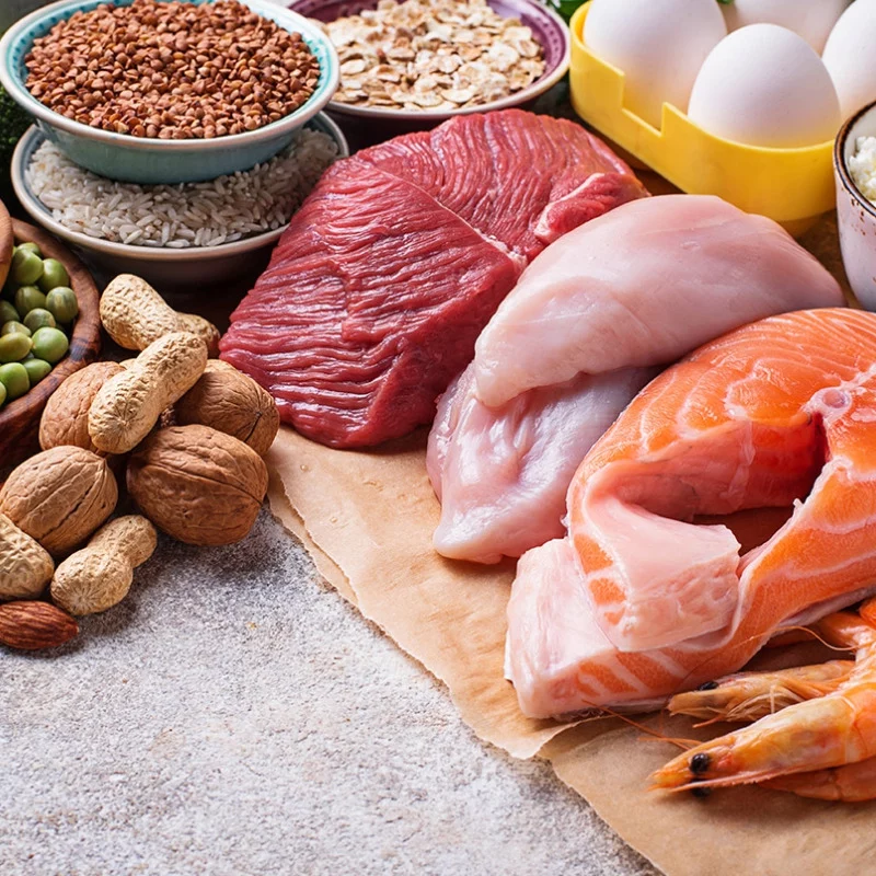 Eine Reihe von eiweißreichen Lebensmitteln, darunter rohes Rindfleisch, Huhn, Lachs, Garnelen, Eier, Erdnüsse, Walnüsse, Edamame, brauner Reis, Hafer und Linsen, auf einem Tisch angeordnet.