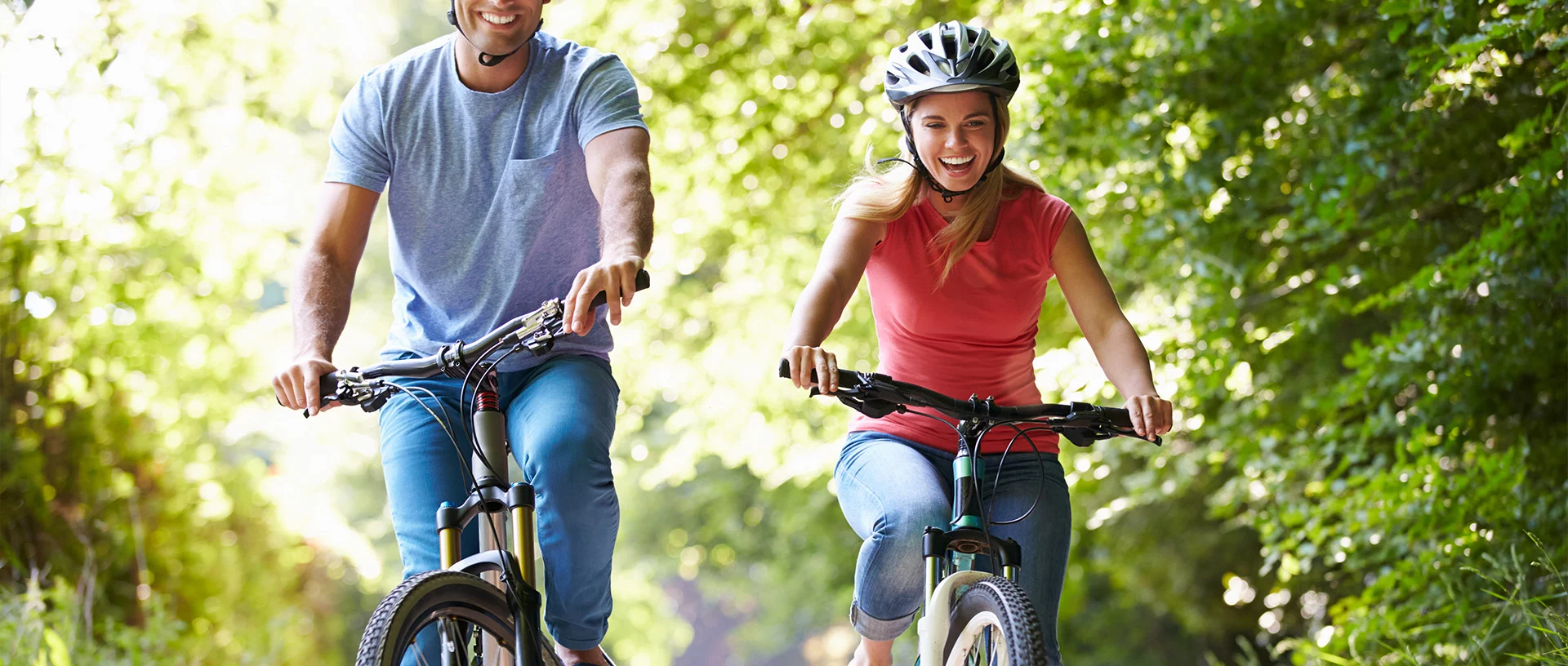 Zwei Personen mit Helmen fahren an einem sonnigen Tag mit dem Fahrrad einen von Bäumen gesäumten Weg entlang. Beide lächeln und genießen die Aktivität im Freien - ein idealer Moment für den Silicium-Blog, um einen lebendigen, gesunden Lebensstil in Aktion festzuhalten.