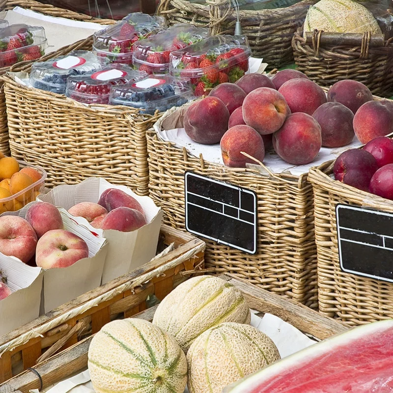 Auf einem Markt im Freien werden Weidenkörbe mit Pfirsichen, Erdbeeren, Melonen und verschiedenen Beeren angeboten. Einige Früchte sind in Kartons und Plastikbehältern verpackt. An den Körben sind Blankoetiketten angebracht.