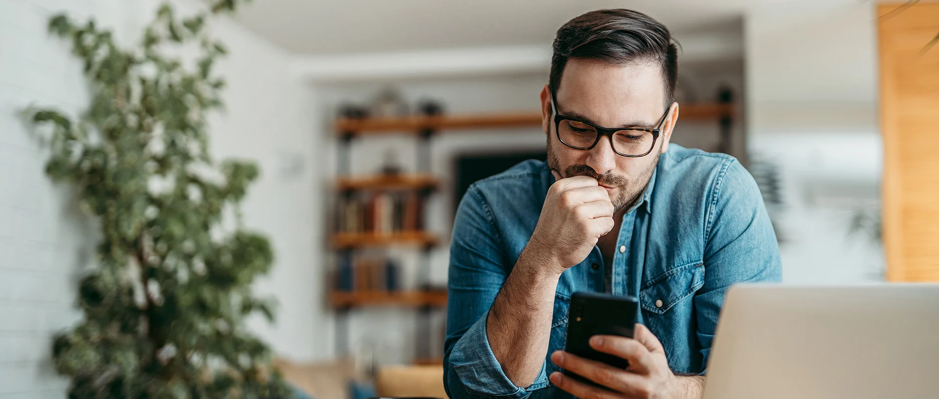 Ein Mann mit Brille und Jeanshemd schaut nachdenklich auf sein Smartphone, während er sich auf einen Tisch stützt. Vor ihm steht ein Laptop, im Hintergrund stehen eine Pflanze und ein Bücherregal.