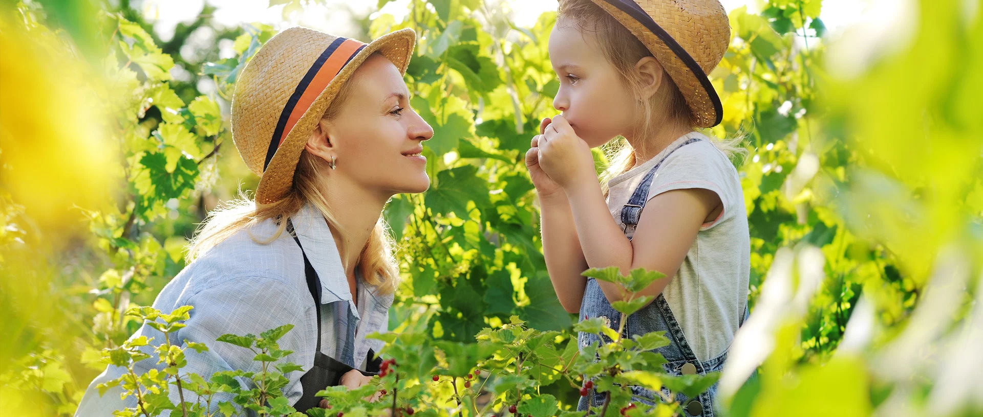 Eine Frau und ein junges Mädchen mit Strohhüten stehen in einem sonnenbeschienenen Garten, umgeben von grünen Pflanzen. Die Frau lächelt das Mädchen an, das sich Beeren an den Mund hält. Beide wirken glücklich und entspannt.
