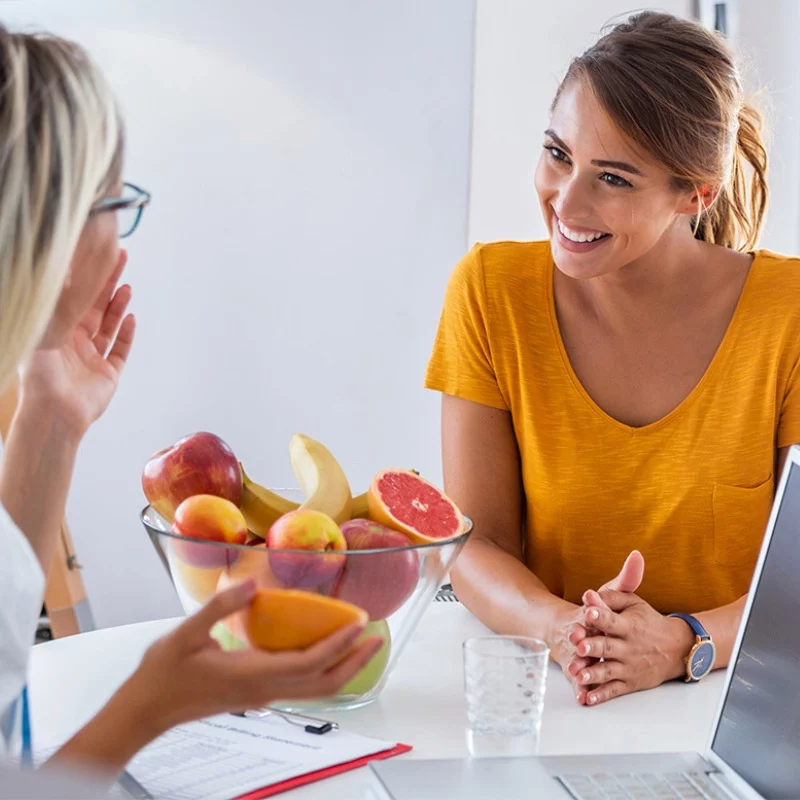 Zwei Frauen sitzen an einem Schreibtisch und führen ein Gespräch. Die eine lächelt und trägt ein gelbes Hemd, während die andere mit einem Stück Obst gestikuliert. Auf dem Tisch zwischen den beiden steht eine Schale mit verschiedenen Früchten, daneben ein Laptop und ein Klemmbrett.