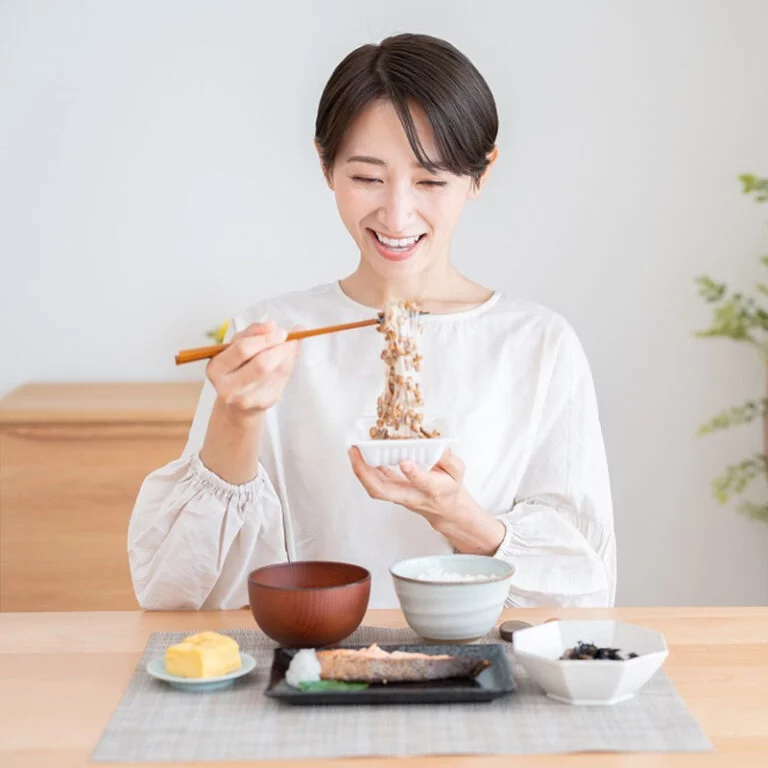 Eine lächelnde Frau sitzt an einem Tisch mit Stäbchen und einer Schüssel und genießt eine traditionelle japanische Mahlzeit mit Gerichten wie Reis, Miso-Suppe, Tamagoyaki, Fisch und Gemüse.
