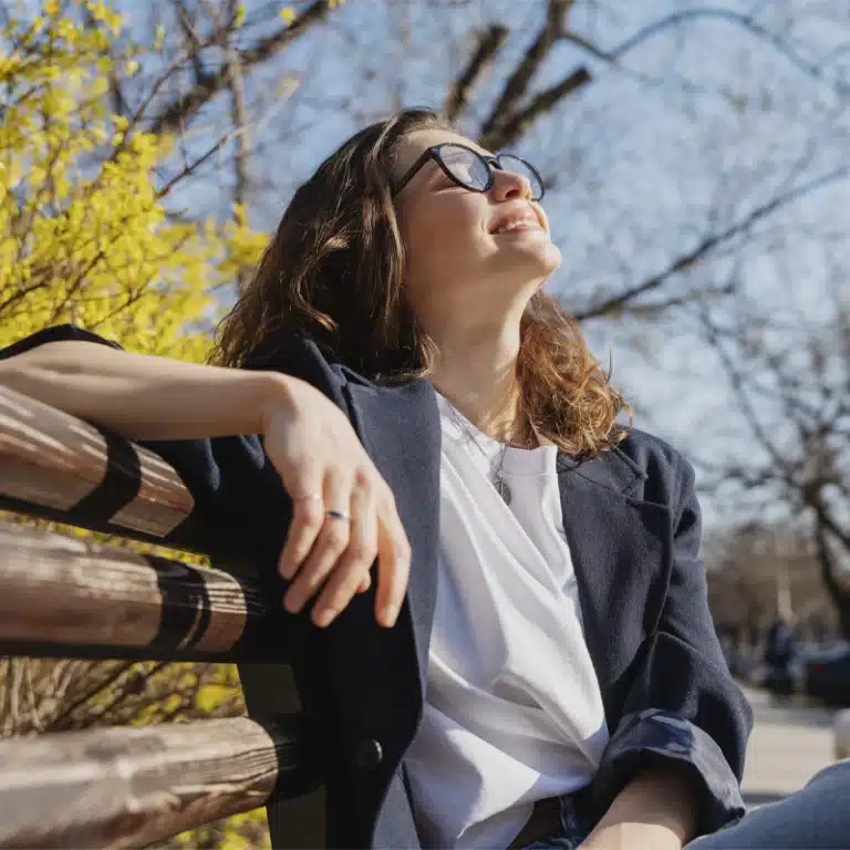 Frau mit Sonnenbrille sitzt auf einer Bank drund streckt ihr Gesicht in die Sonne