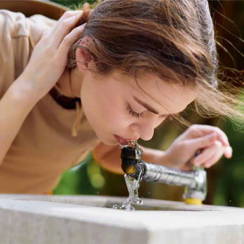 Eine Frau, die sich mit einer Hand die Haare aus dem Gesicht hält, trinkt Wasser aus einem Brunnen im Freien. Der Brunnen sieht aus wie ein rechteckiges Waschbecken aus Beton.