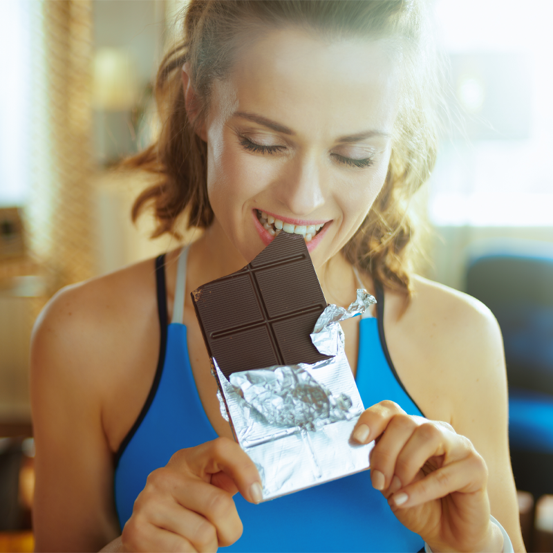 Eine sportliche Frau in einem blauen Fitness-Top beißt genussvoll in eine Tafel dunkle Schokolade.