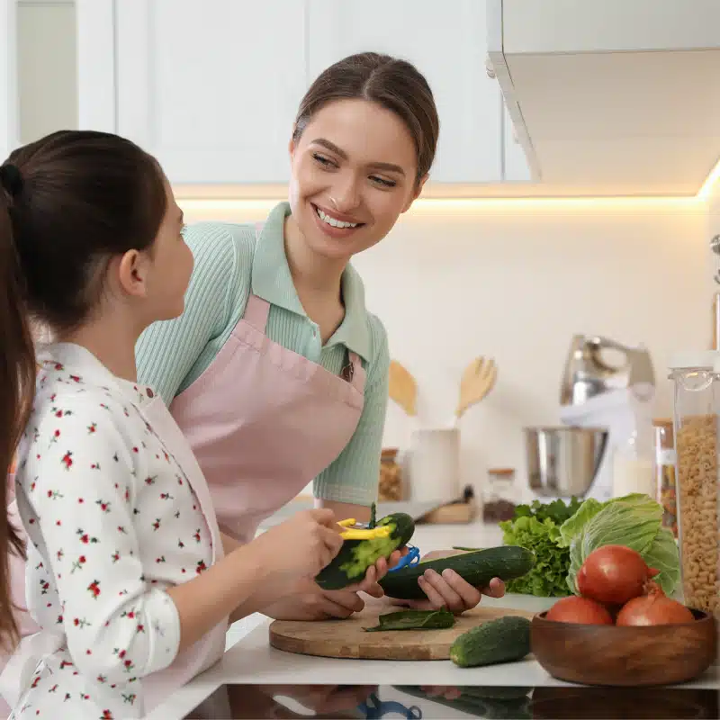 Mutter und Tochter bereiten in der Küche gemeinsam frische Zutaten vor: Zwei Personen schneiden und schälen Gemüse für die Mahlzeitenplanung mit Gurken, Salat, Kohl und Tomaten