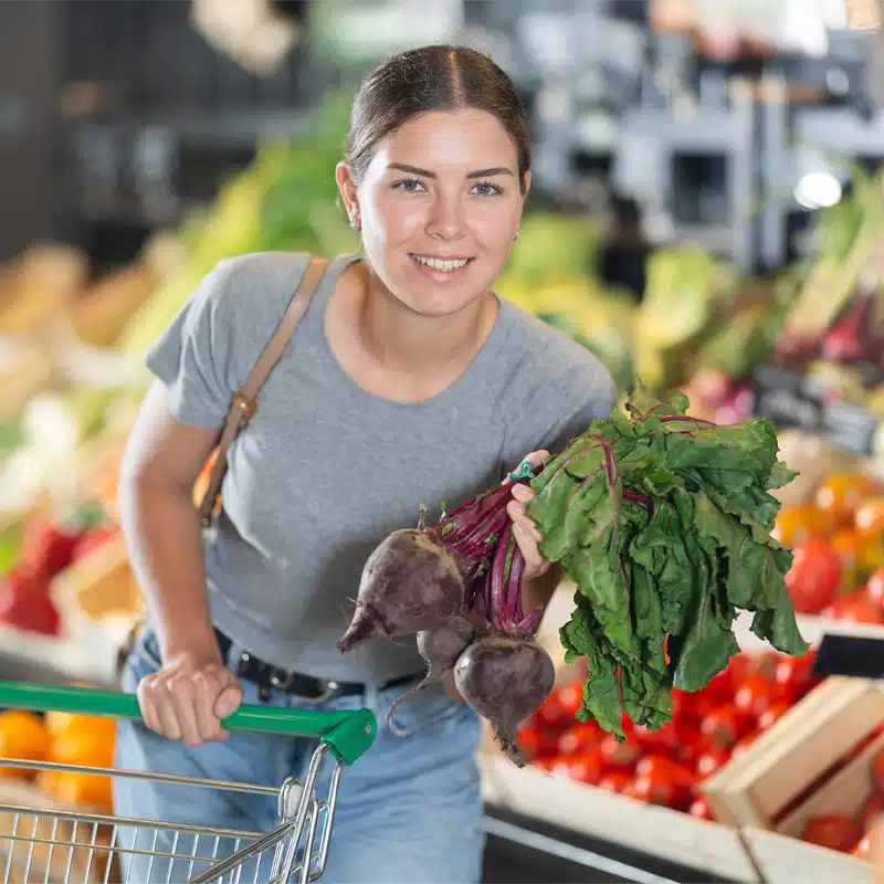 Frau mit Einkaufswagen hält frische Rote Bete auf einem Gemüsemarkt vor einer Auslage mit regionalem Obst und Gemüse.