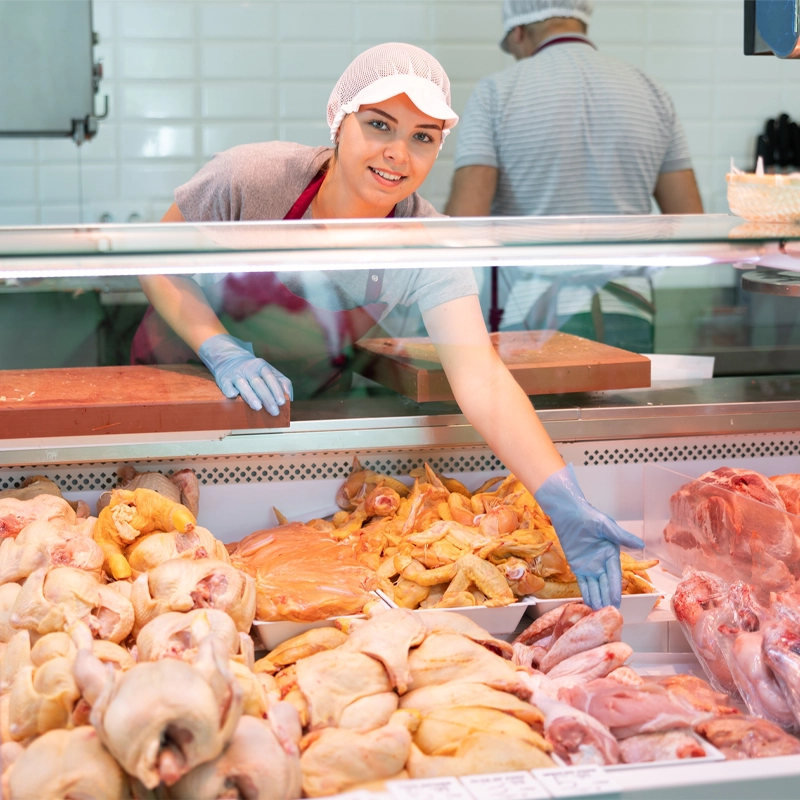 Frau mit Hygieneschutzkleidung verkauft frisches Geflügelfleisch an der Fleischtheke eines Lebensmittelgeschäfts.